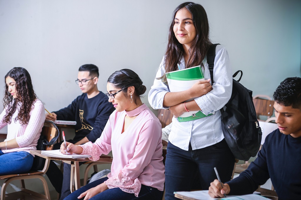 A girl holding a book and standing in the center of a classroom.